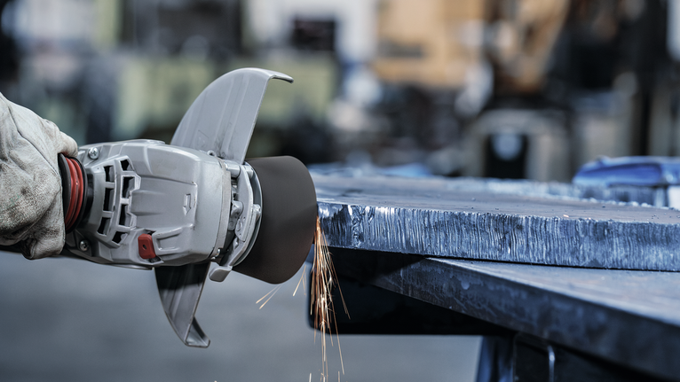 A person wearing safety equipment grinds the edge of a large metal sheet.