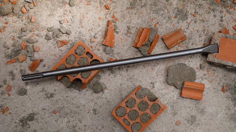 Chisel bit lying on a concrete surface surrounded by broken bricks.