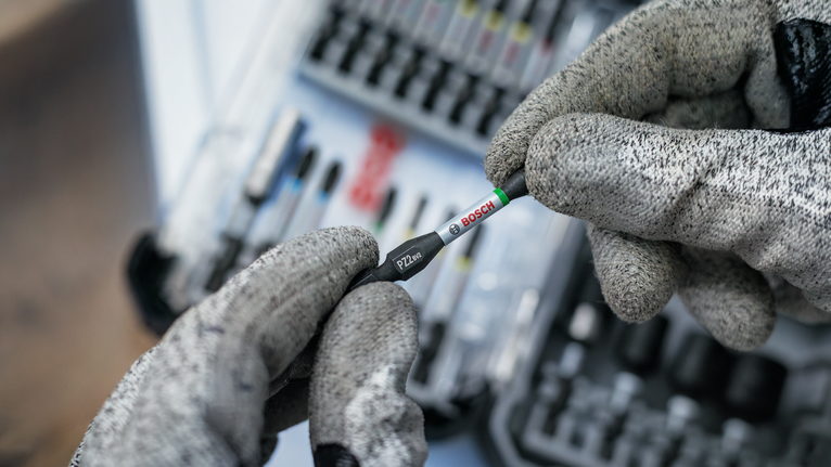 Person wearing safety equipment holds a screwdriver bit near a tool kit.