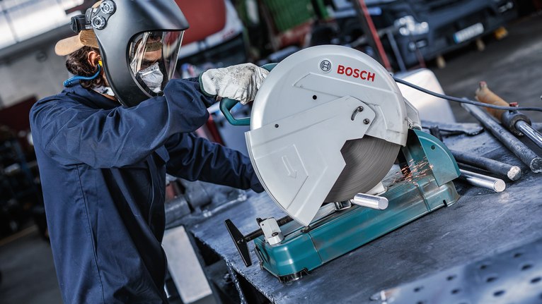 A person wearing safety equipment cuts metal pipes with a stationary circular saw.