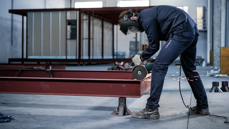 A person wearing safety equipment grinds metal beams in an industrial workshop.