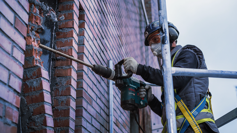 A person wearing safety equipment drills into a brick wall using a power tool.