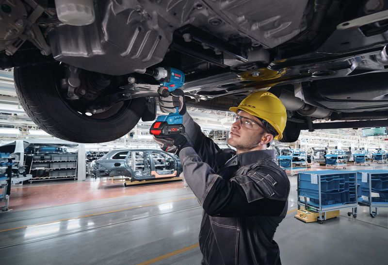 A person wearing safety equipment uses a power wrench to fasten bolts under a vehicle chassis.