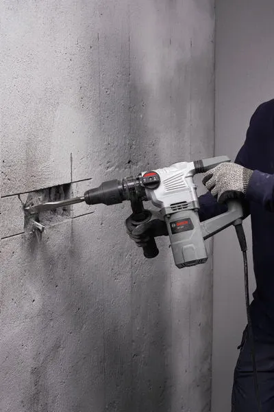 A person wearing safety equipment uses a rotary hammer to chisel a groove in a concrete wall.