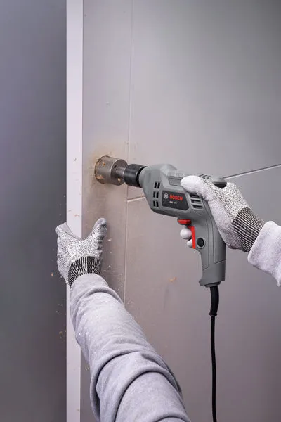 A person wearing safety equipment uses a drill to make a hole in a wall.