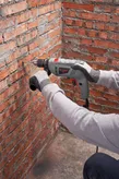 A person wearing safety equipment drills into a red brick wall with a power tool.