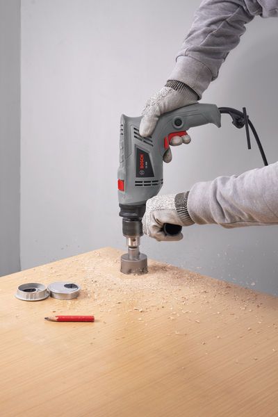 A person wearing safety equipment drills a large hole in a wooden board.