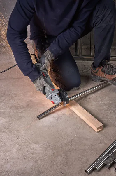 A person wearing safety equipment cuts a metal pipe with a grinder on a concrete floor.