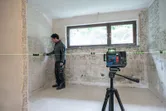 A person marks tiles on a wall using a laser leveling tool on a tripod.