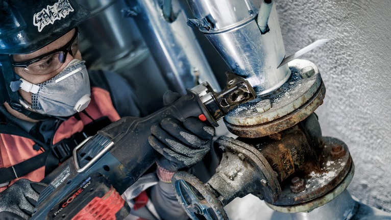 A person wearing safety equipment uses a reciprocating saw on a rusty pipe joint.
