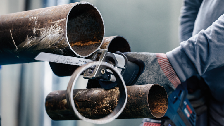 Person wearing safety equipment cuts a metal pipe with a reciprocating saw.