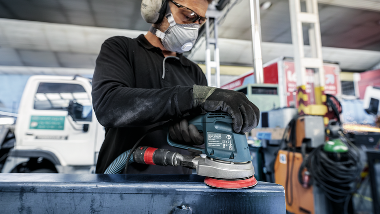 Person wearing safety equipment sands a metal surface with a power sander.