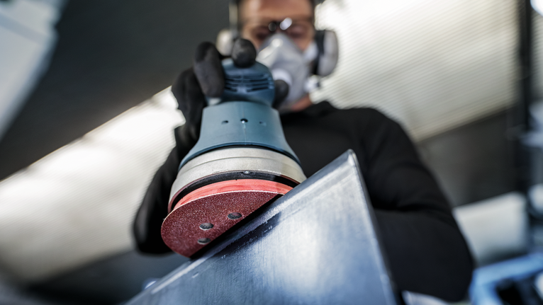 Person wearing safety equipment uses a random orbital sander on a metal surface.