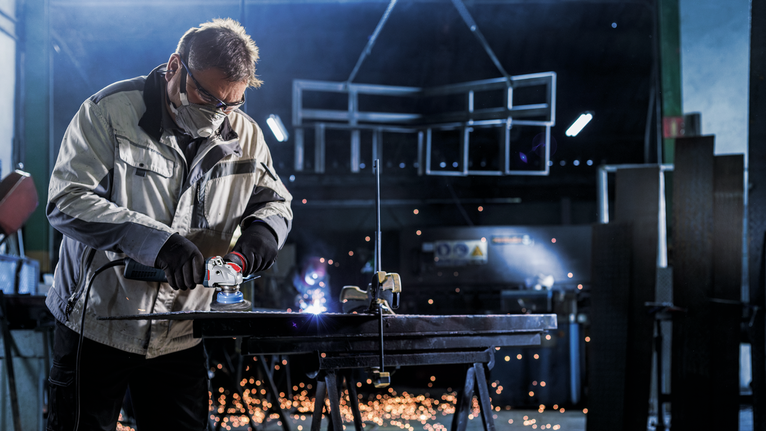 A person wearing safety equipment uses a grinder on metal, with sparks flying in a workshop.