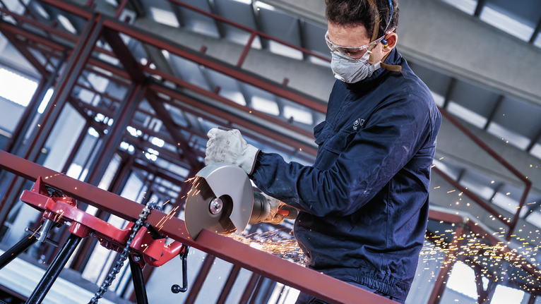 A person wearing safety equipment uses a power cutter to cut a metal pipe, producing sparks.