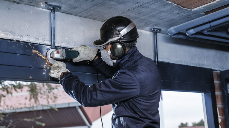 Person wearing safety equipment grinds metal beams, producing sparks.