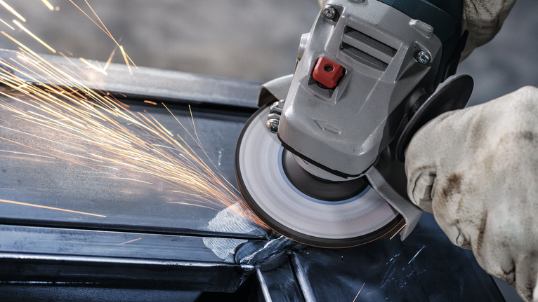 Person wearing safety equipment grinds a metal beam with an angle grinder.