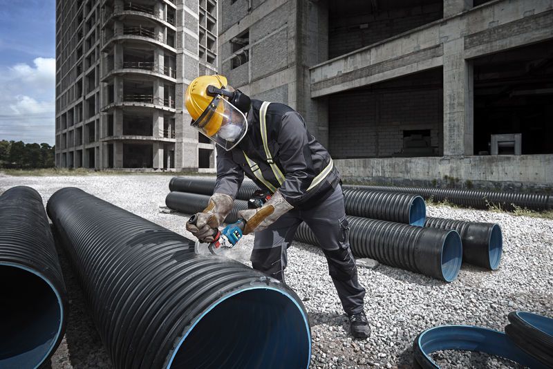 A person wearing safety equipment cuts a large plastic pipe with a cordless angle grinder.