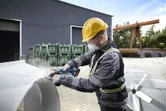 A person wearing safety equipment grinds metal outdoors at an industrial site.
