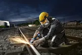 A person wearing safety equipment uses an angle grinder to cut a railway track.