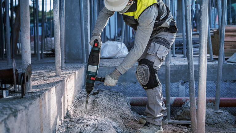 A person wearing safety equipment uses a power tool to break concrete.