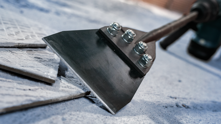 Wide chisel attachment removing ceramic tiles from a concrete floor.