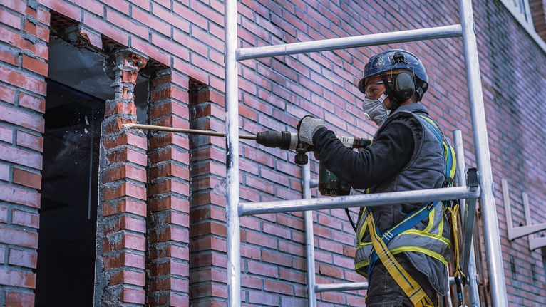 A person wearing safety equipment drills into a brick wall.