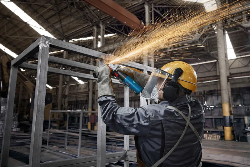 A person wearing safety equipment grinds metal, creating sparks in a factory.