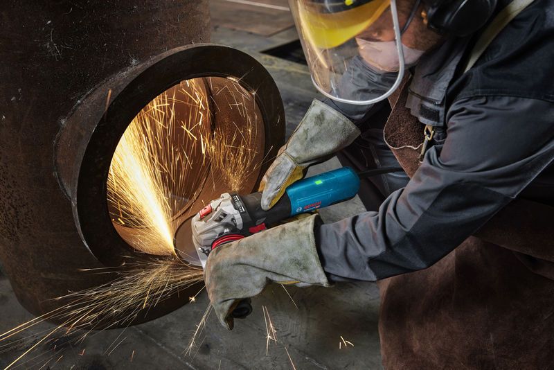 A person wearing safety equipment grinds a large metal pipe, creating bright sparks.