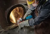A person wearing safety equipment grinds a large metal pipe, creating bright sparks.