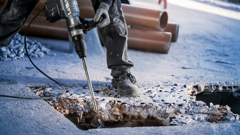 Person wearing safety equipment breaks concrete floor with a heavy-duty power tool.