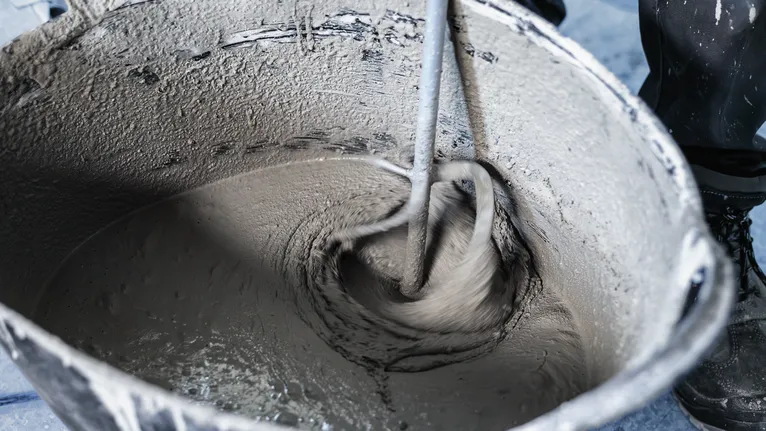 A person wearing safety equipment mixes cement with a power tool in a bucket.