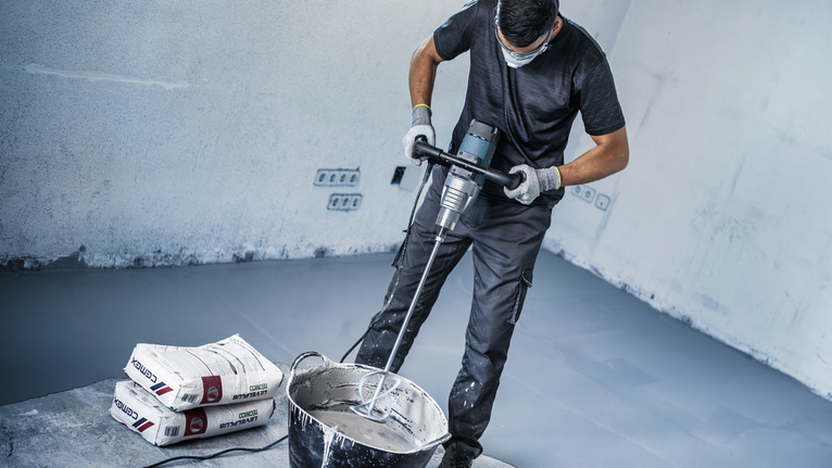 Person wearing safety equipment mixes cement in a bucket with a power mixer.