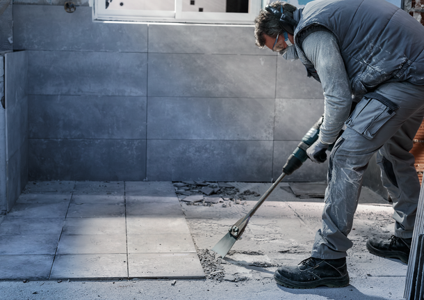 Person wearing safety equipment removes floor tiles with a demolition tool.