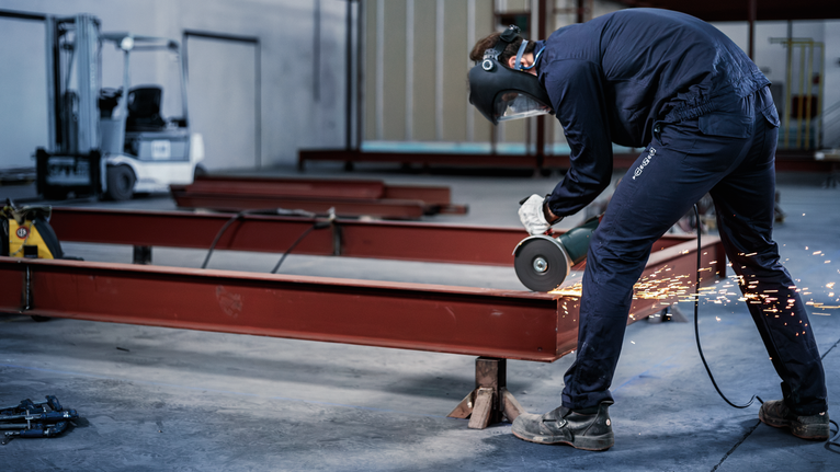A person wearing safety equipment grinds a metal beam, sparks flying.