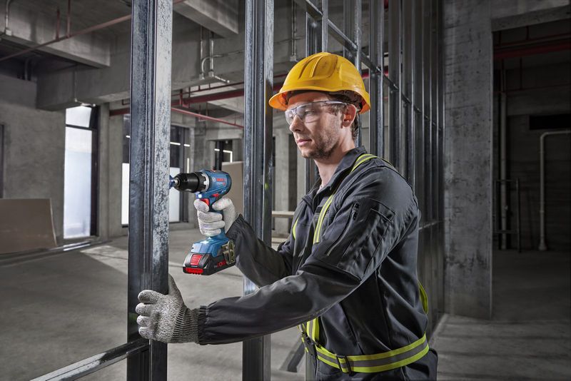 A person wearing safety equipment uses a cordless drill on a metal frame.