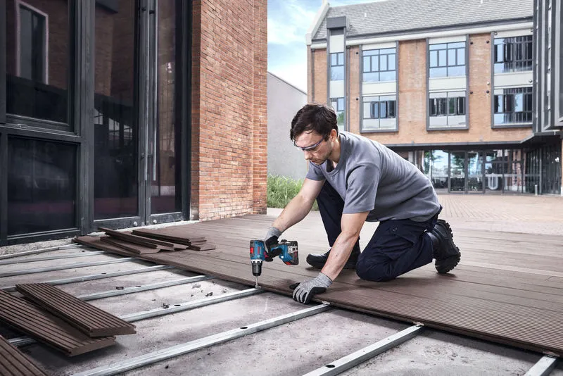 A person wearing safety equipment uses a cordless drill to install decking boards outside.
