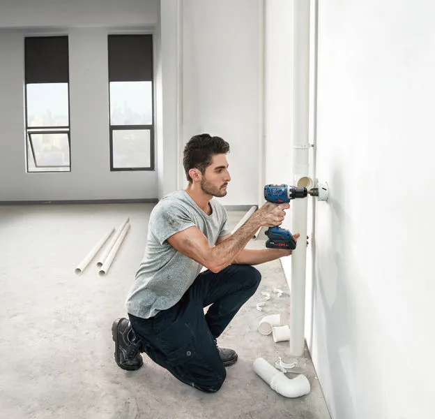 A person drills into a wall beside plumbing pipes in a partially finished room.