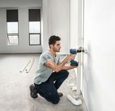 A person drills into a wall beside plumbing pipes in a partially finished room.