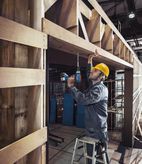 A person wearing safety equipment drills into a wooden beam while standing on a ladder.