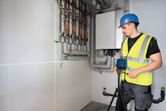 A person wearing safety equipment sets up a laser leveling tool near wall pipes.
