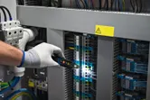 A person wearing safety equipment checks electrical wiring in a control panel with a voltage tester.