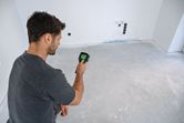 A person scans a bare room floor, using a handheld thermo camera.