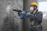 A person wearing safety equipment uses a demolition hammer to break a concrete wall.