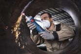 A person wearing safety equipment grinds the inside of a large metal pipe.