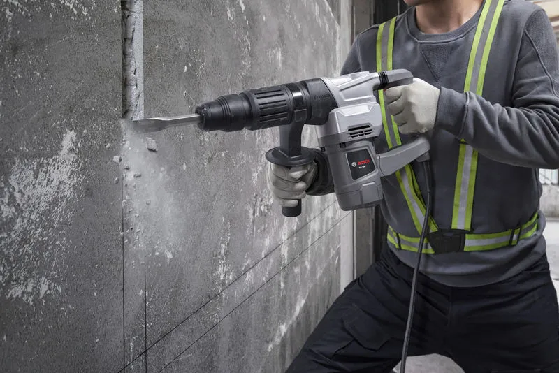 A person wearing safety equipment uses a demolition hammer to break concrete wall.