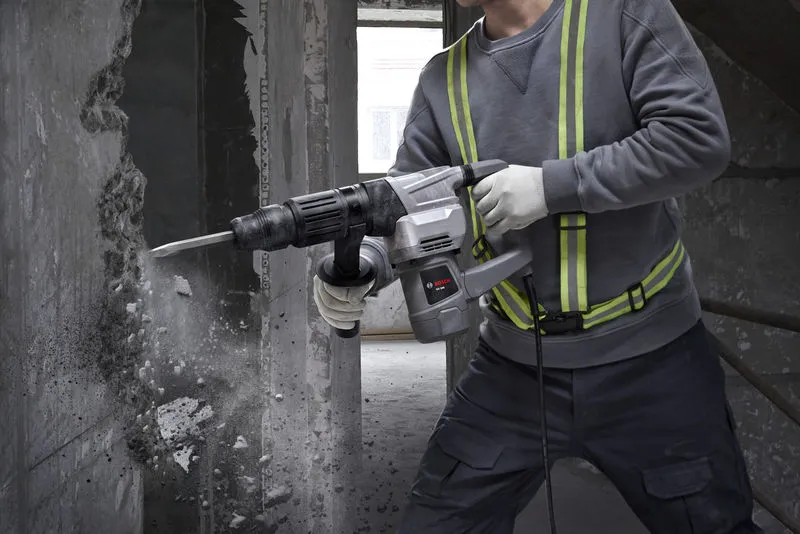 A person wearing safety equipment uses a demolition hammer to break a concrete wall.