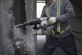A person wearing safety equipment uses a demolition hammer to break a concrete wall.