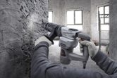 A person wearing safety equipment uses a demolition hammer to break a concrete wall.