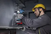 A person wearing safety equipment tightens bolts on a steel beam using a cordless impact wrench.